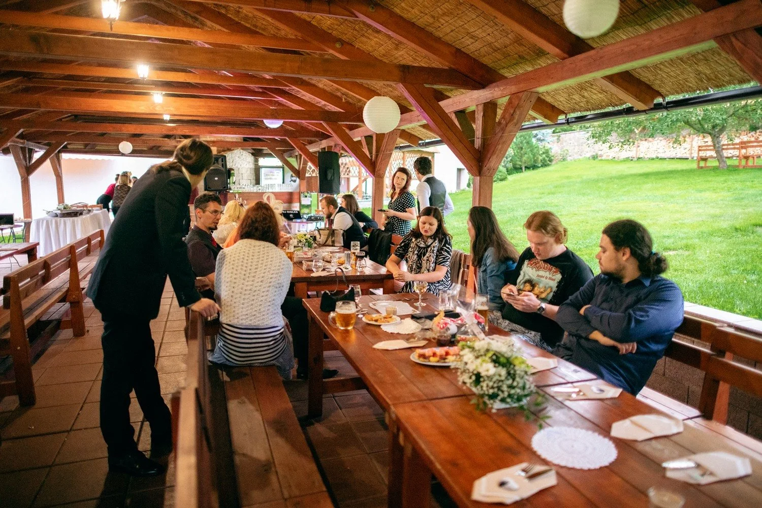 Večerní hostina pokračuje u dlouhého stolu v pavilonu.
