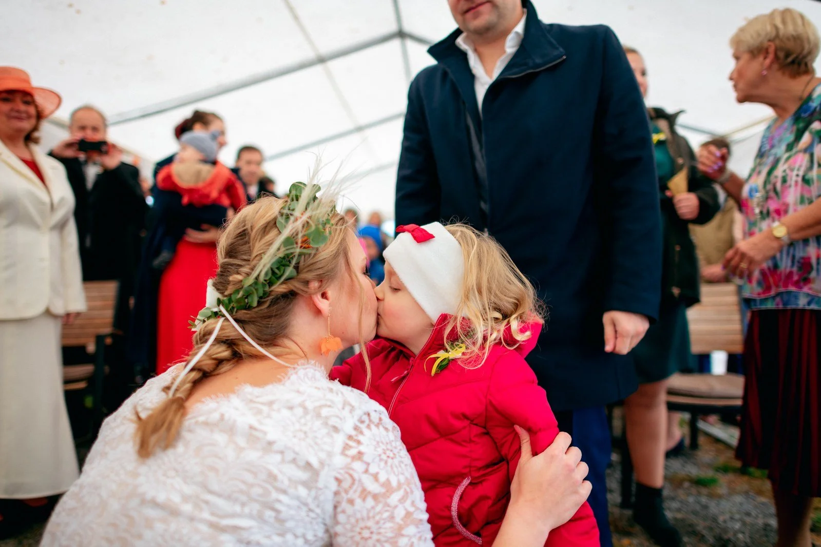 Žena v bílých šatech klečí před malou dívkou v červeném oděvu mezi hosty.