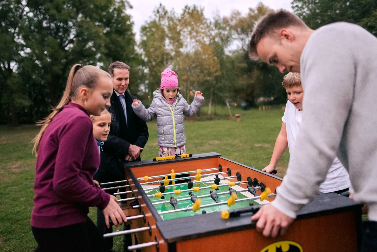 Čtyři lidé hrají venku stolní fotbal, další hosté stojí v pozadí.