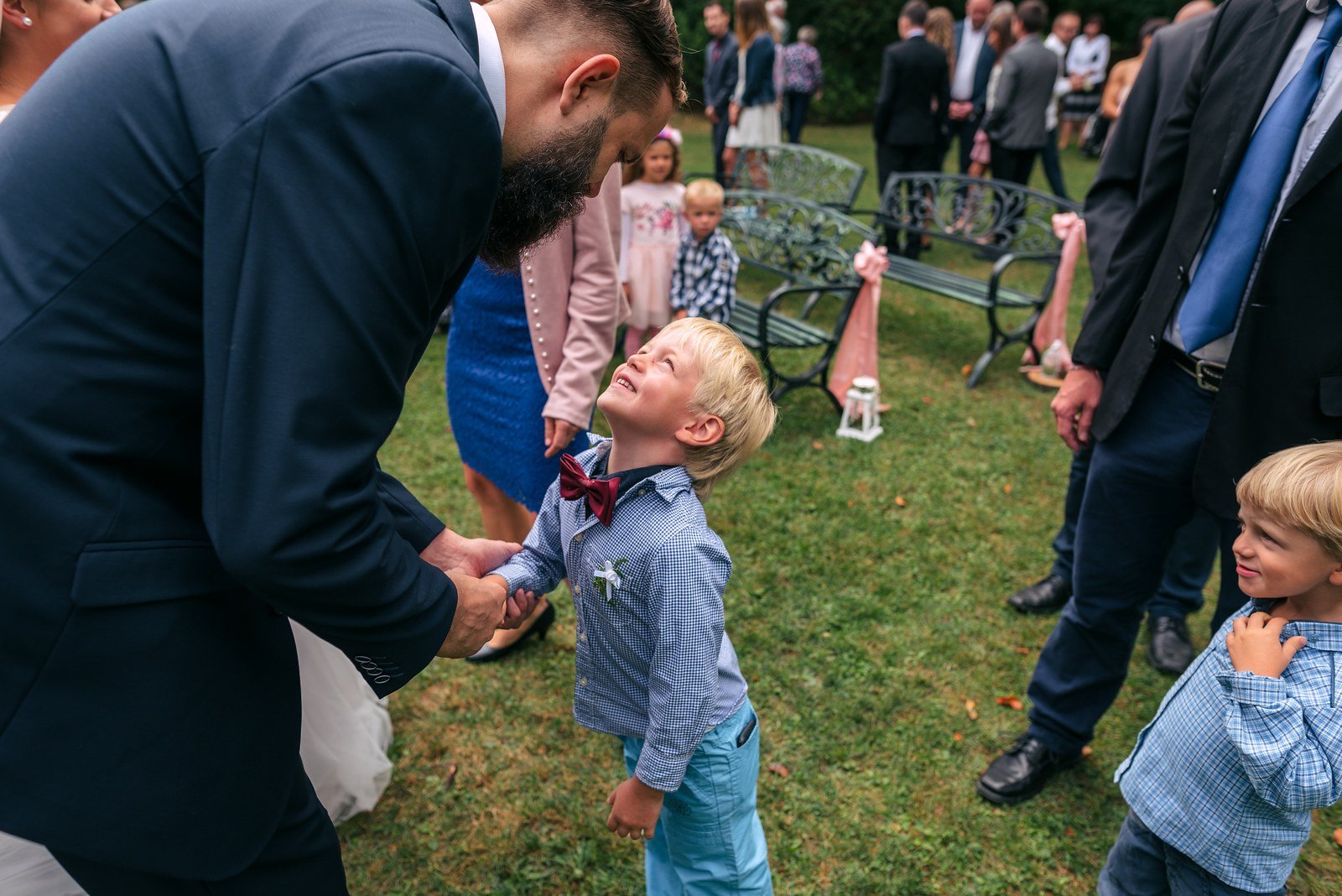 Muž v tmavém obleku se sklání k malému chlapci uprostřed skupiny hostů.
