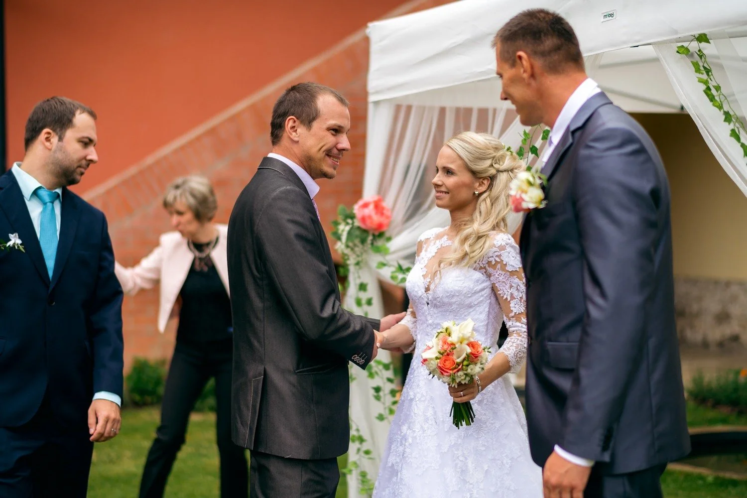 Nevěsta si podává ruku s mužem v tmavém obleku, zatímco ženich stojí po jejím boku.