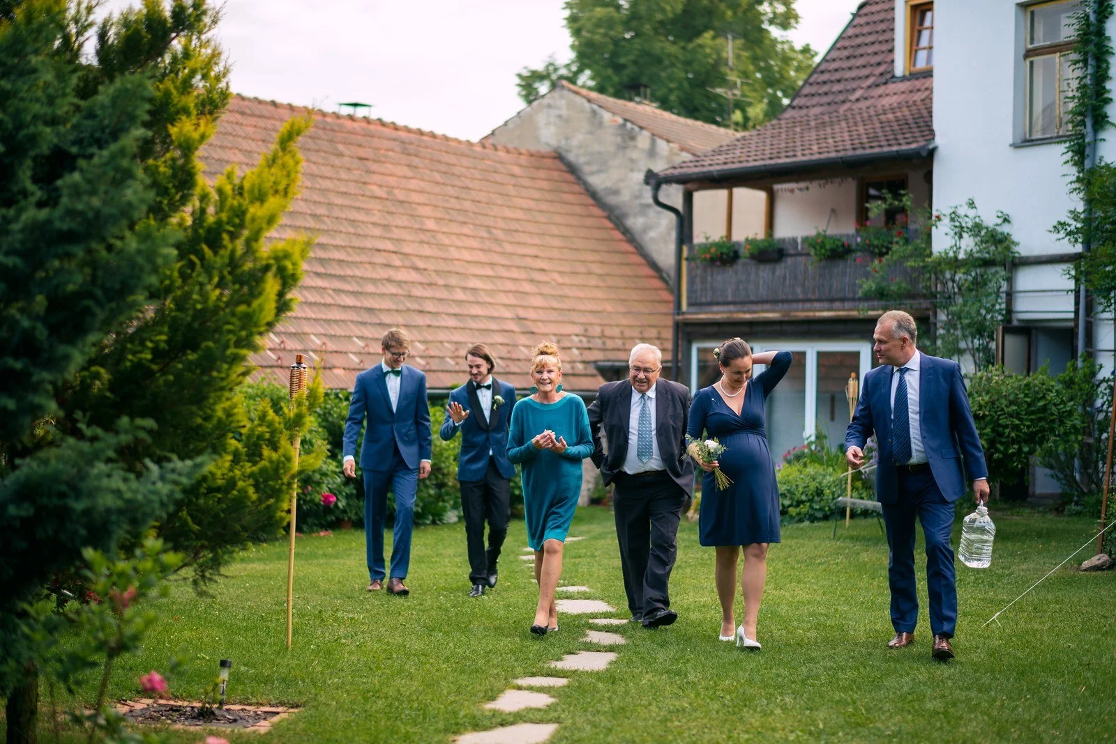Skupina slavnostně oblečených lidí přichází po kamenné pěšině zahradou k domu.