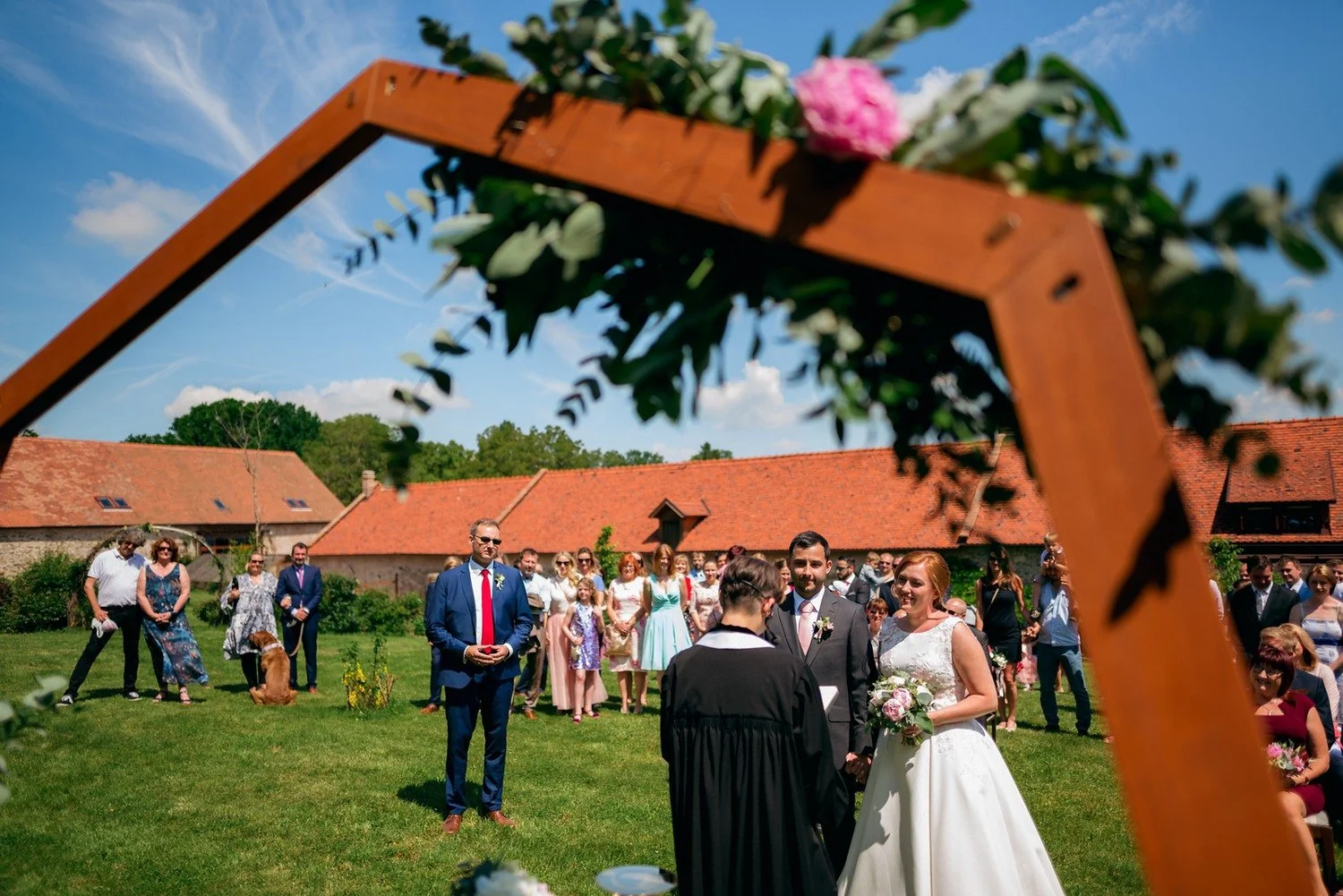 Nevěsta se ženichem jsou při obřadu zachyceni skrz dřevěný rám zdobený zelení a růžovou květinou.