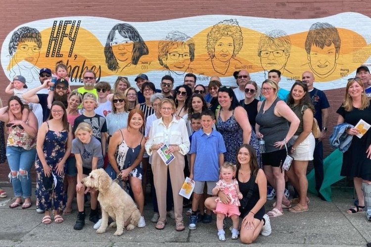 A group of 30+ people of all ages pose in front of a mural on a wall in Lawrenceville. The mural shows six older women and the text inscription reads: "Hey, babes."