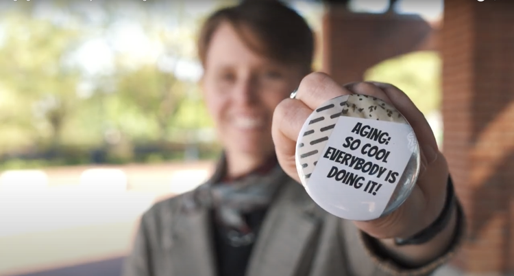 A blurred out shot of Christine Happel holding a button which reads Aging: So Cool Everybody Is Doing It!
