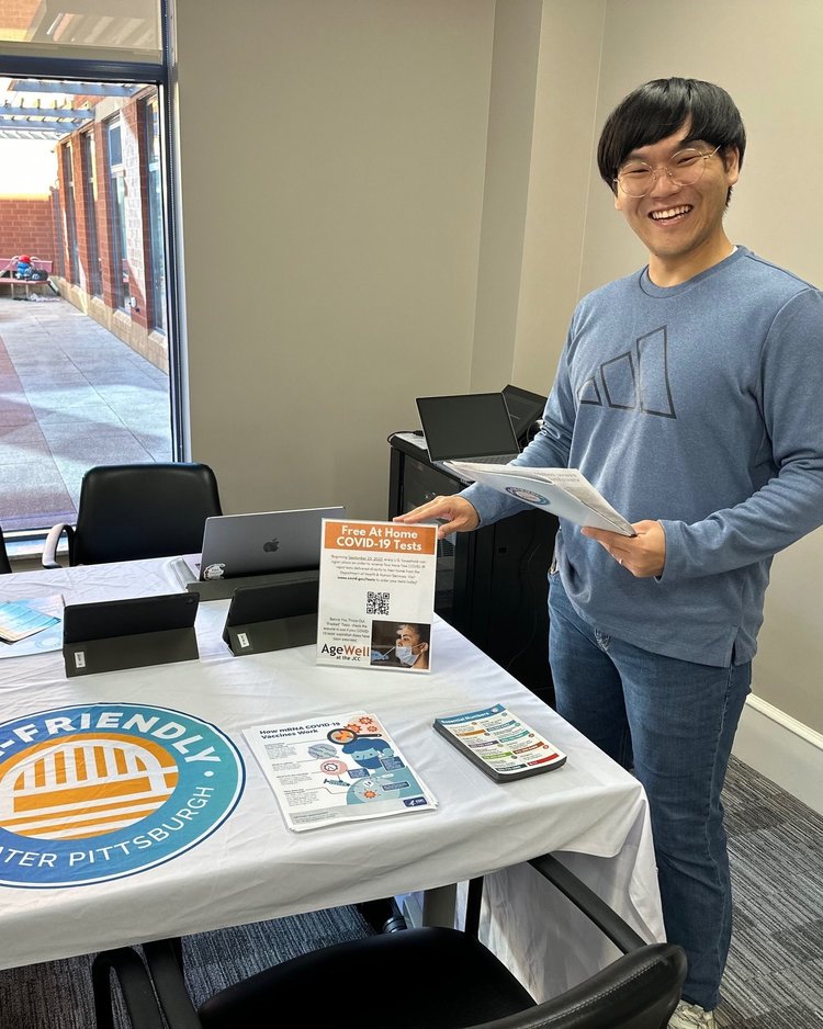 A younger Korean man smiles and poses before a table with the Age-Friendly Greater Pittsburgh logo. 