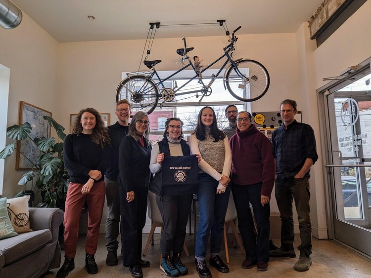 Eight people pose for a group photo in front of a bike hanging from the ceiling. One of the people is holding an Age-Friendly Greater Pittsburgh tote bag.