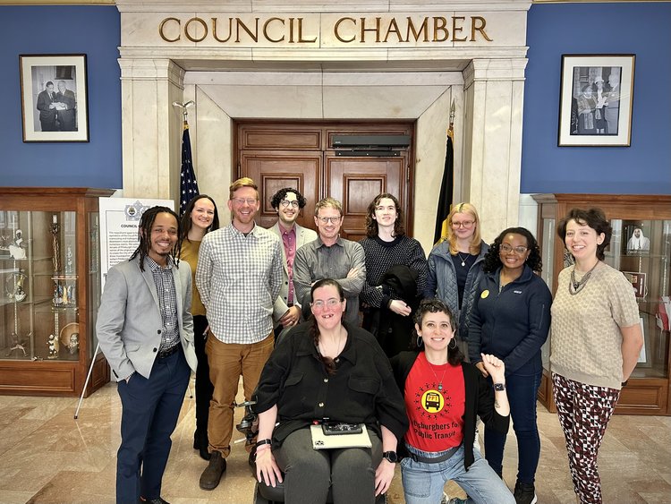 Eleven people of all races, genders and ages pose for a group photo beneath the entrance of the Pittsburgh City Council Chamber.