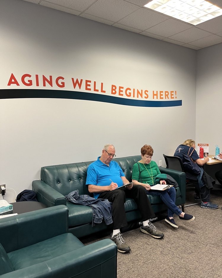 Two people sit on couches and fill out forms under a sign that says "Aging Well Beings Here!"