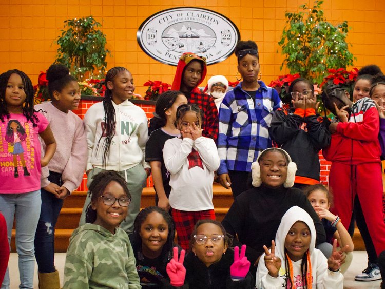 About 15 students pose for a group photo in front of an orange wall that is decorated with Christmas trees and features the Clairton city seal.