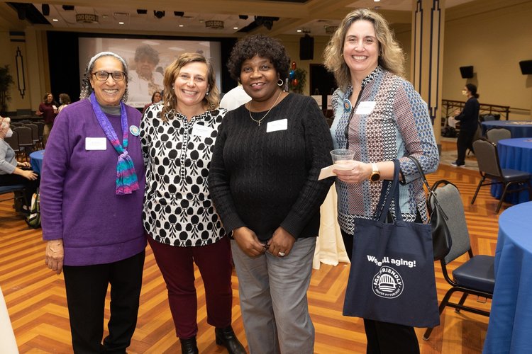 Four women smile and pose for group photo. One of the women is holding a tote bag with the Age-Friendly Greater Pittsburgh logo and the phrase "We're All Aging"!