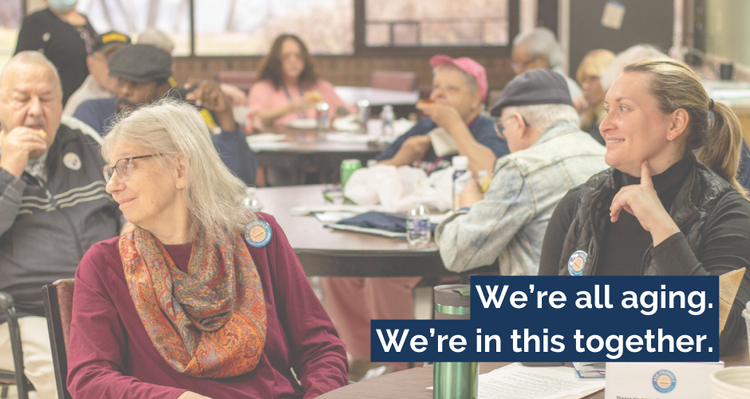 A photograph of about 20 people in a lunchroom smiling and socializing. The text reads: "We're all aging. We're all in this together."