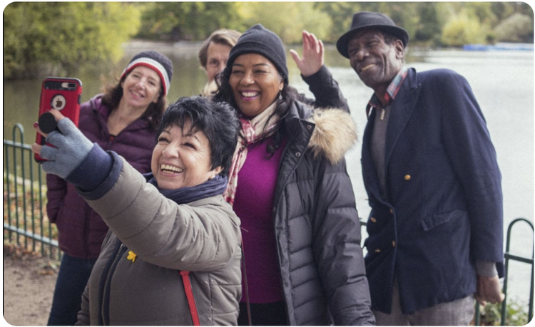 Five people of all races, genders and ages take a selfie in a park by the pond.