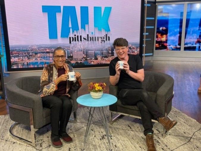 An older woman and a younger man sit in nice chairs and sip coffee on the set of Talk Pittsburgh.