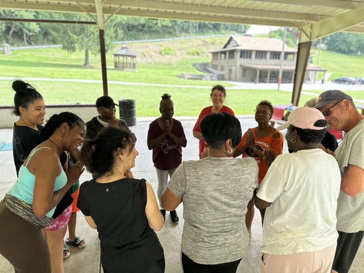 Eleven people of all ages stand in a circle under a park pavilion. They are wearing yoga clothes and enjoying a moment of calm reflection.