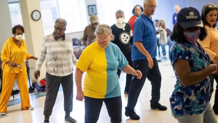 9 older adults dance in a brightly lit room.