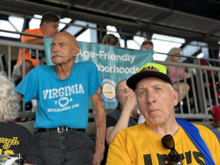 10+ people of all ages are watching a baseball game. Many people are wearing black and yellow Pirates jerseys. In the background there is a banner with the words "Age-Friendly Neighborhoods" and the Age-Friendly Greater Pittsburgh logo.