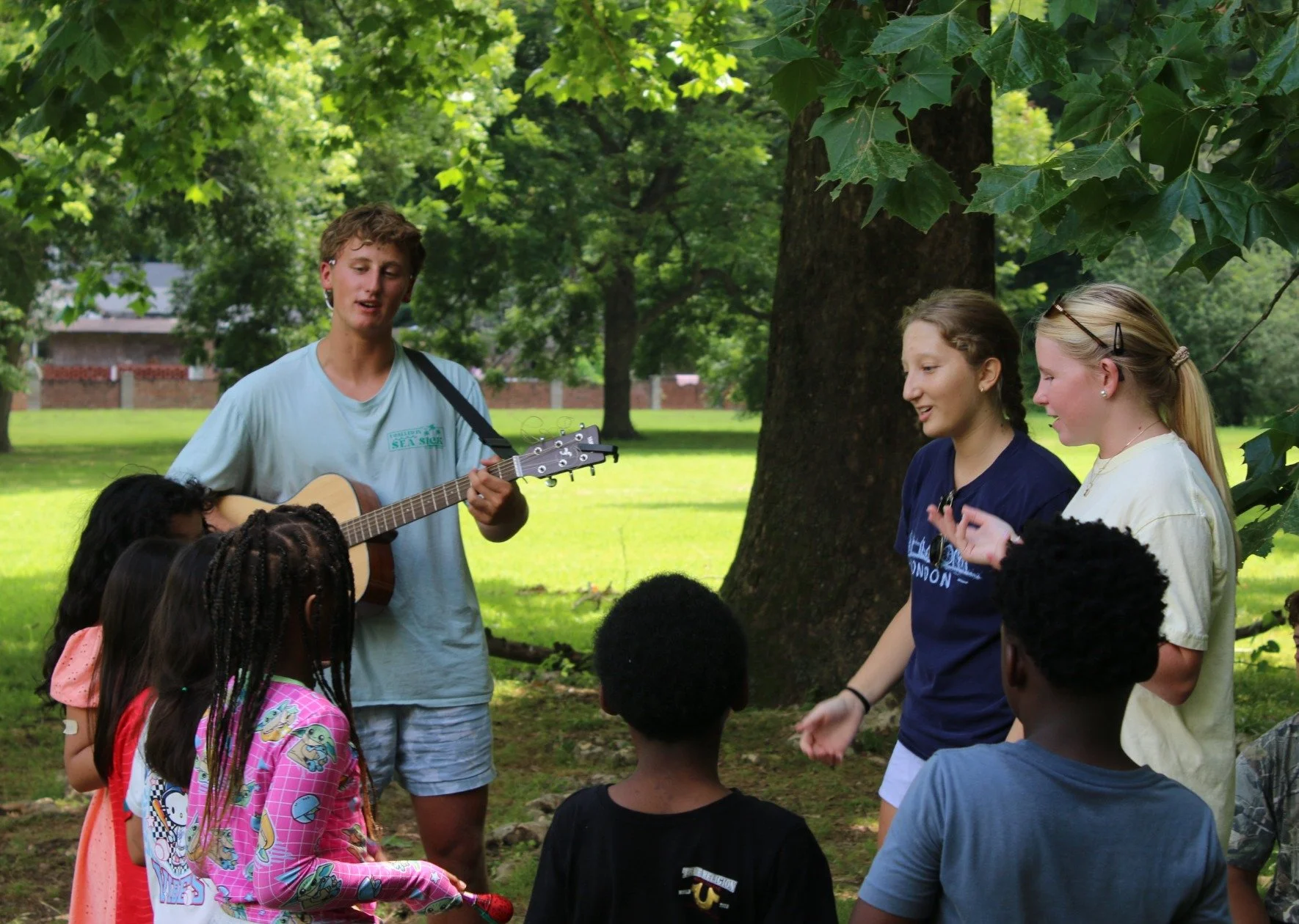 Young people leading singing with a guitar as children gather outdoors under a tree.