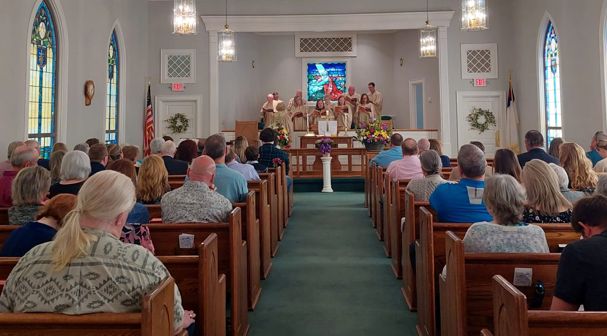 A congregation seated in wooden pews as a choir sings at the front of a sanctuary with stained-glass windows.