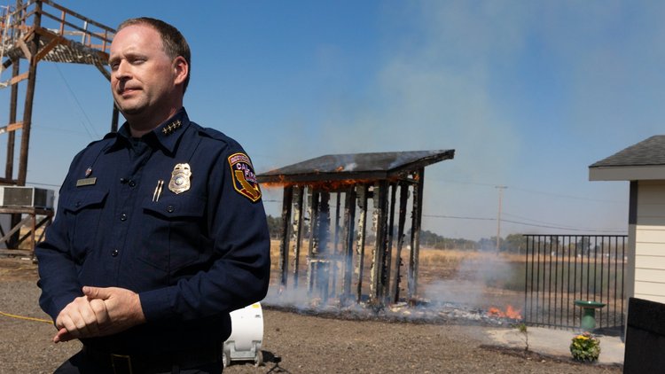 At the site of a fire safety demonstration, a firefighter stands in front of smoldering structure and part of a surviving structure. 