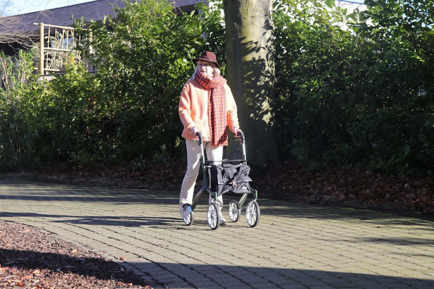 Older woman walking on a paved path with a four-wheeled walker while looking to her left.