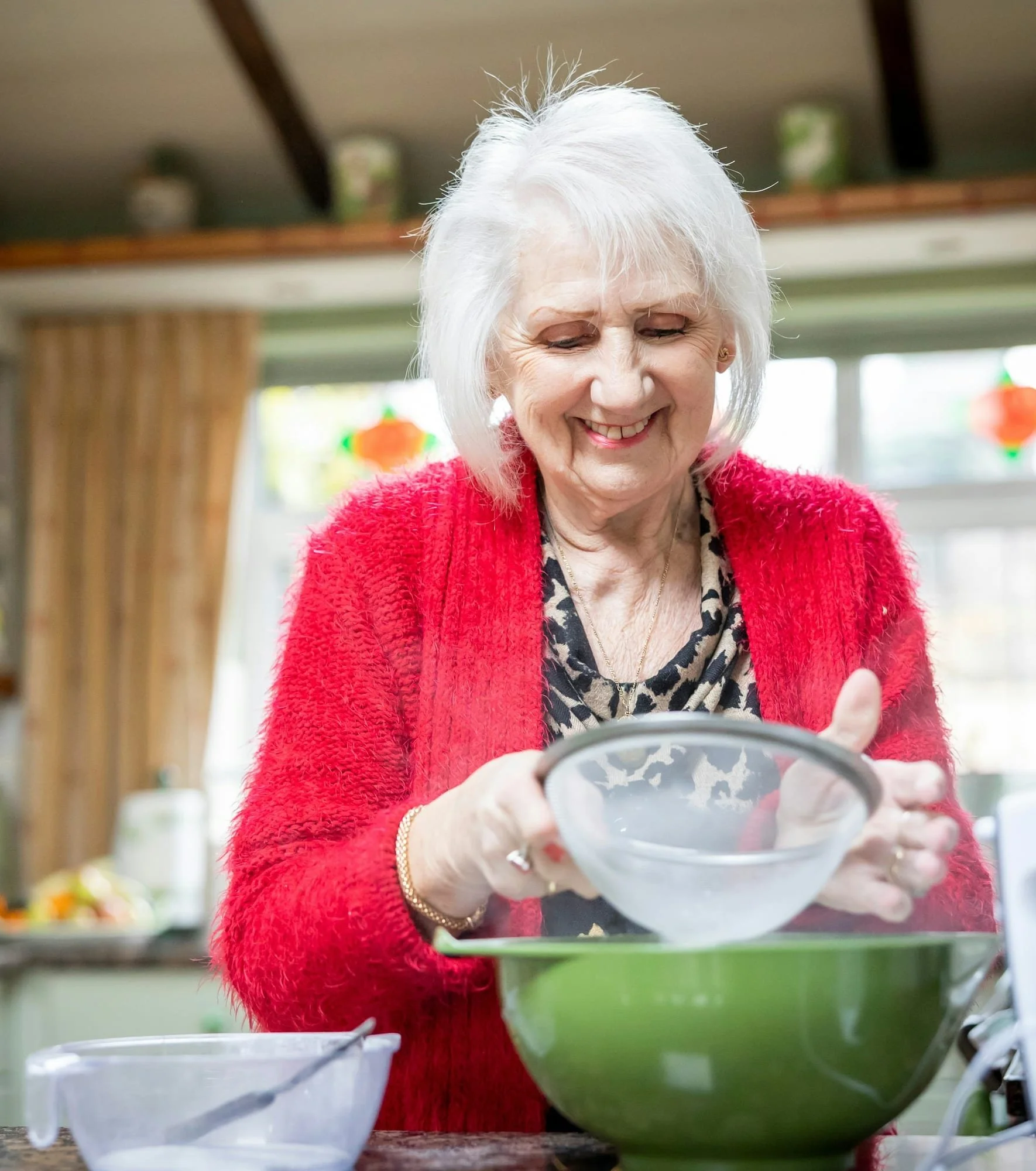 Older woman sifting flour into a mixing bowl in her kitchen.