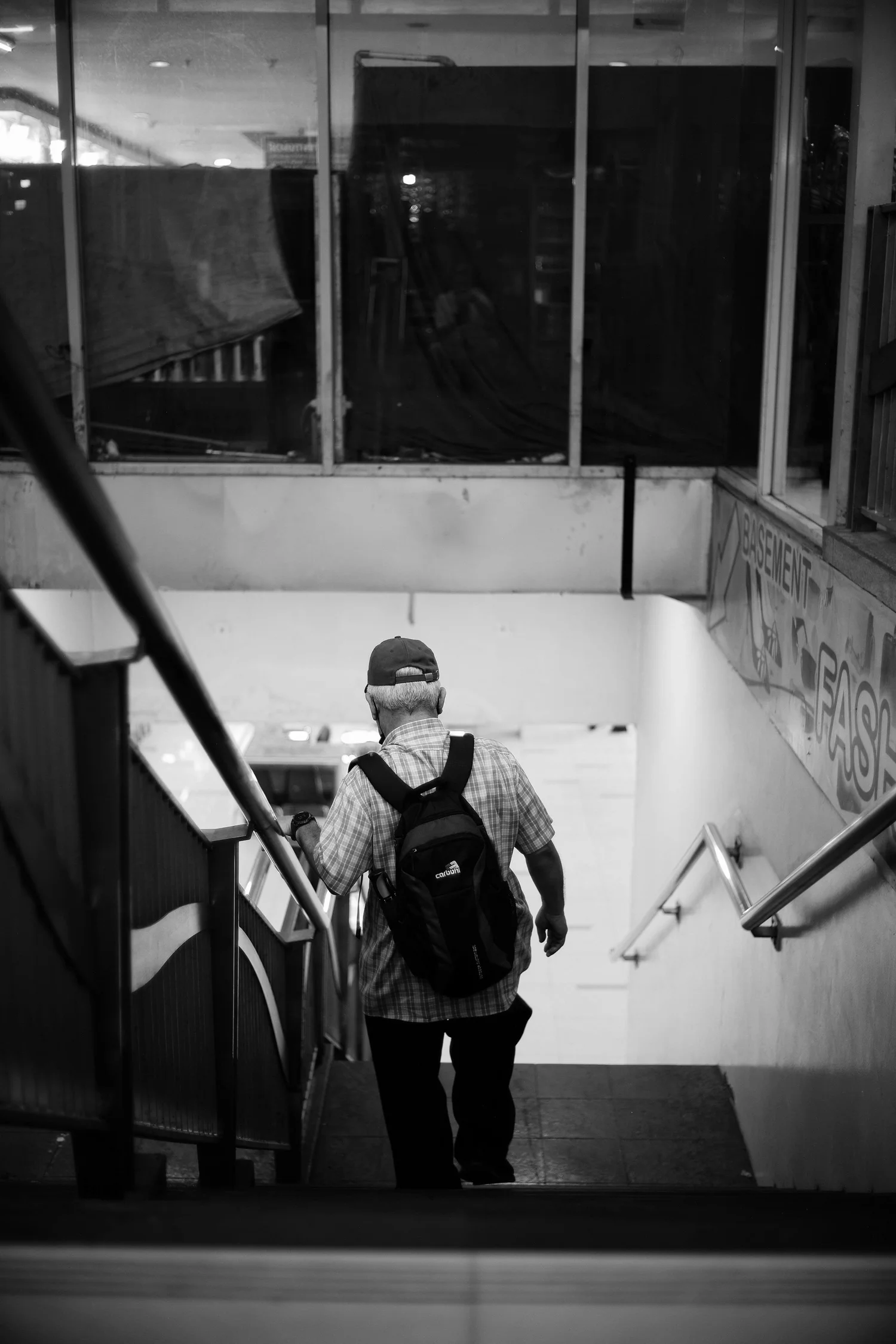 Older man with a backpack walks down an indoor staircase beside a handrail.