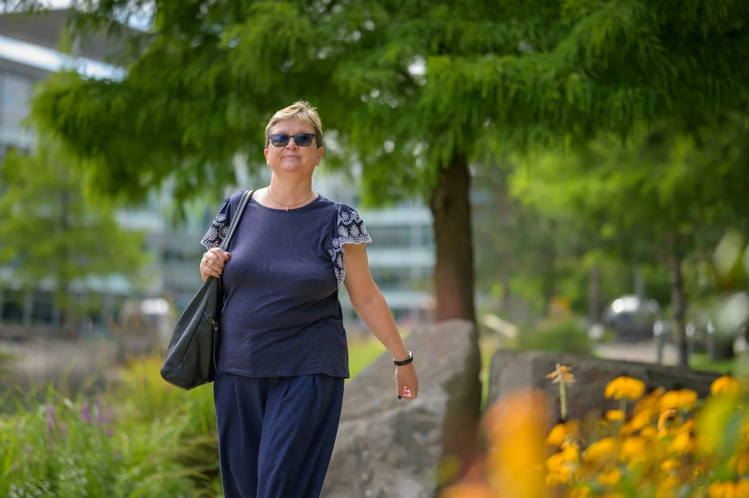 Older woman walking along a landscaped path carrying a shoulder bag.