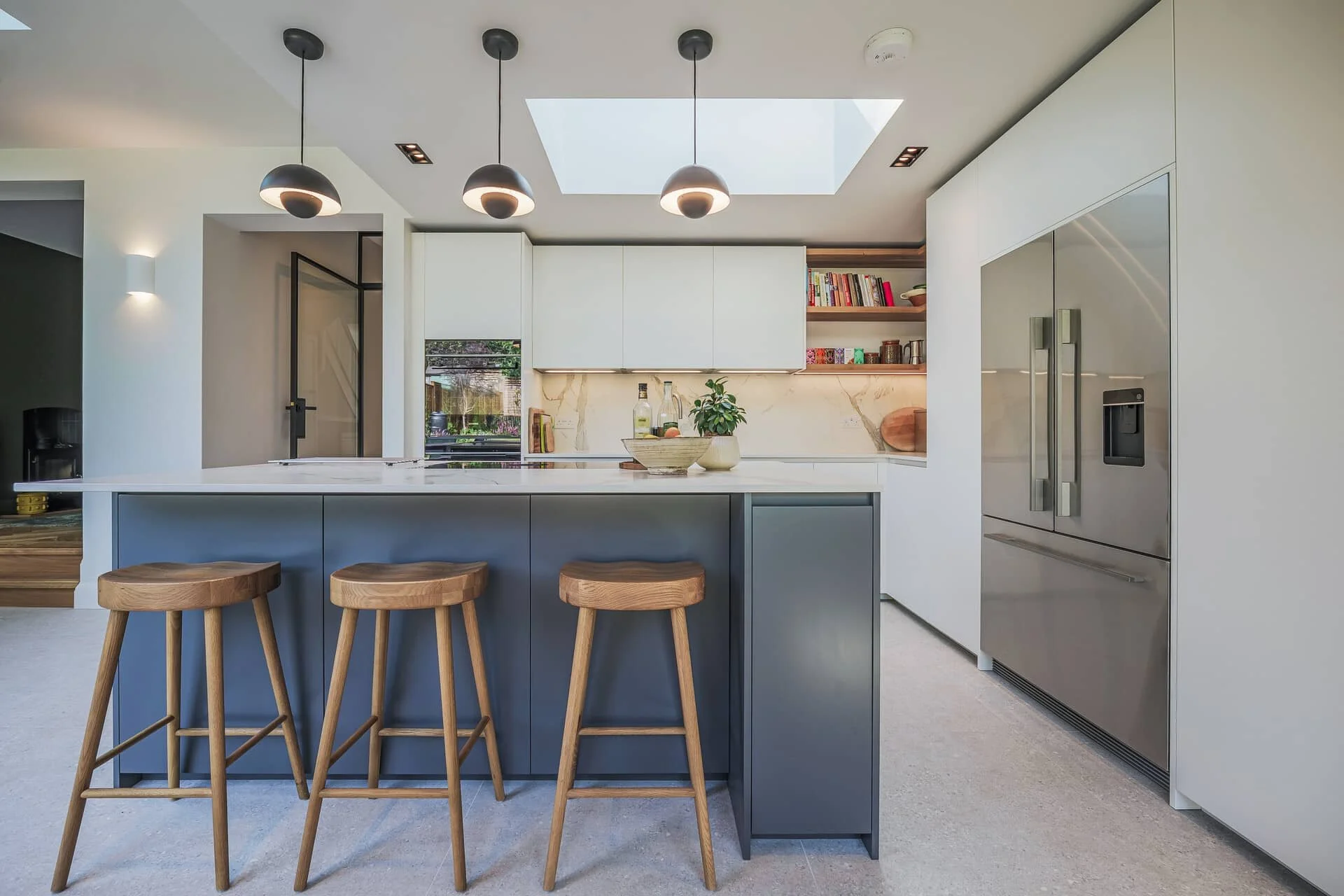 Open-plan kitchen with a slate blue island under three black dome pendants and a skylight, white worktop and cabinetry, a veined marble splashback behind the hob, open timber shelving holding books and jars, and a flush-fitted American-style fridge freezer.