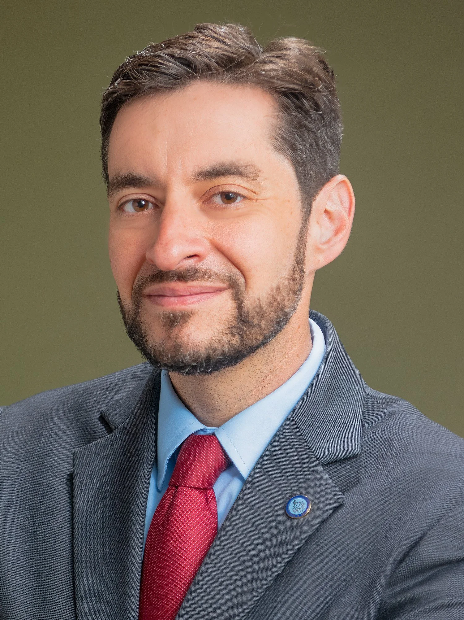 Headshot of a smiling bearded man in a gray suit, light blue shirt, red tie and lapel pin against an olive green backdrop
