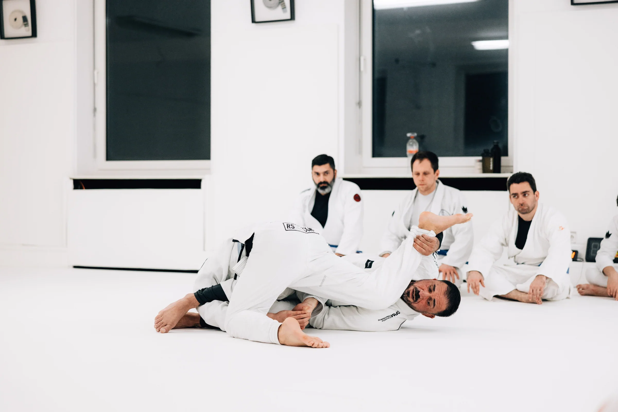 Zwei Menschen zeigen eine BJJ-Bodensequenz, die Trainingsgruppe schaut zu