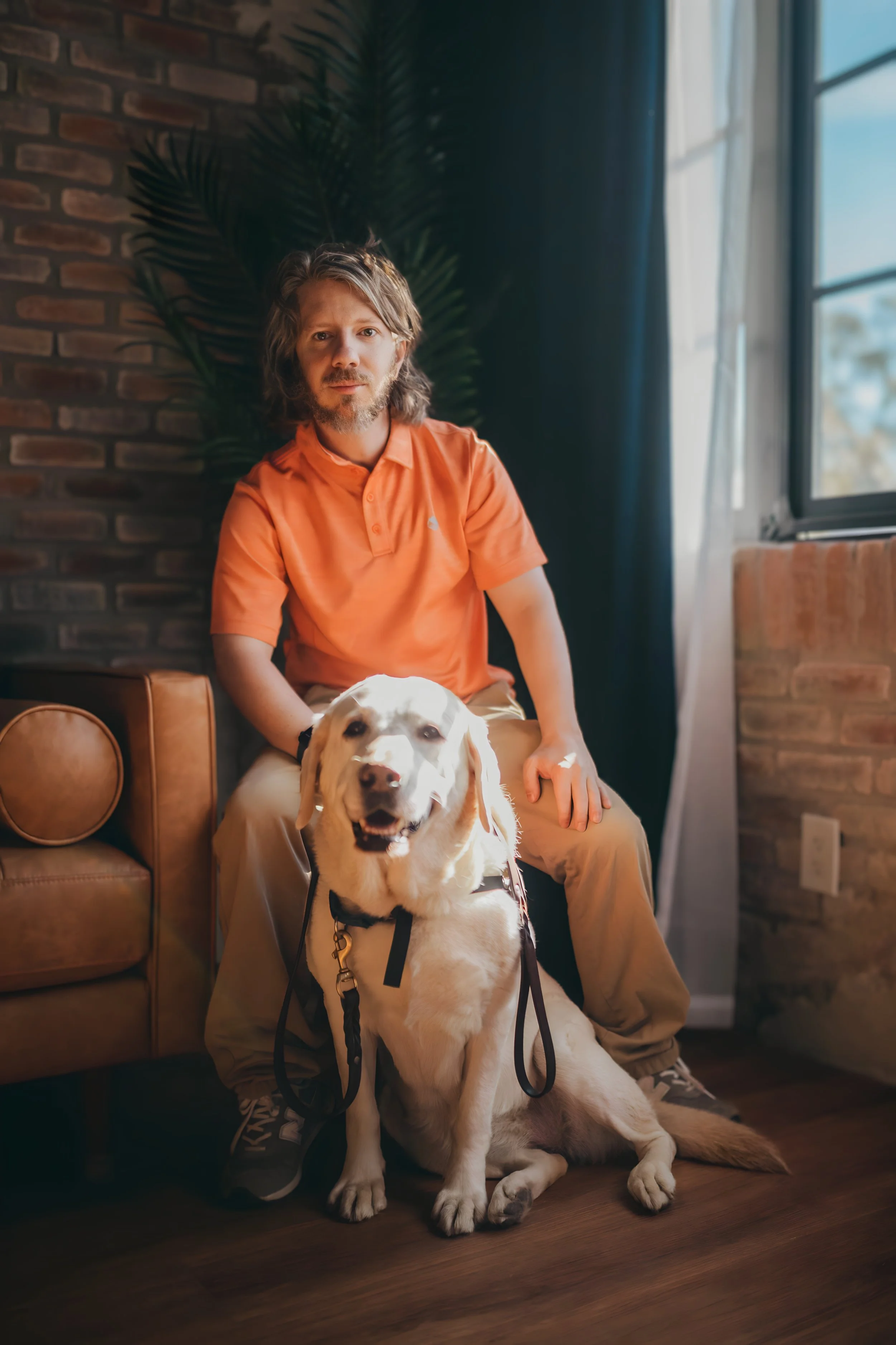 Brendan Oakes seated on a leather couch beside his yellow Labrador service dog.