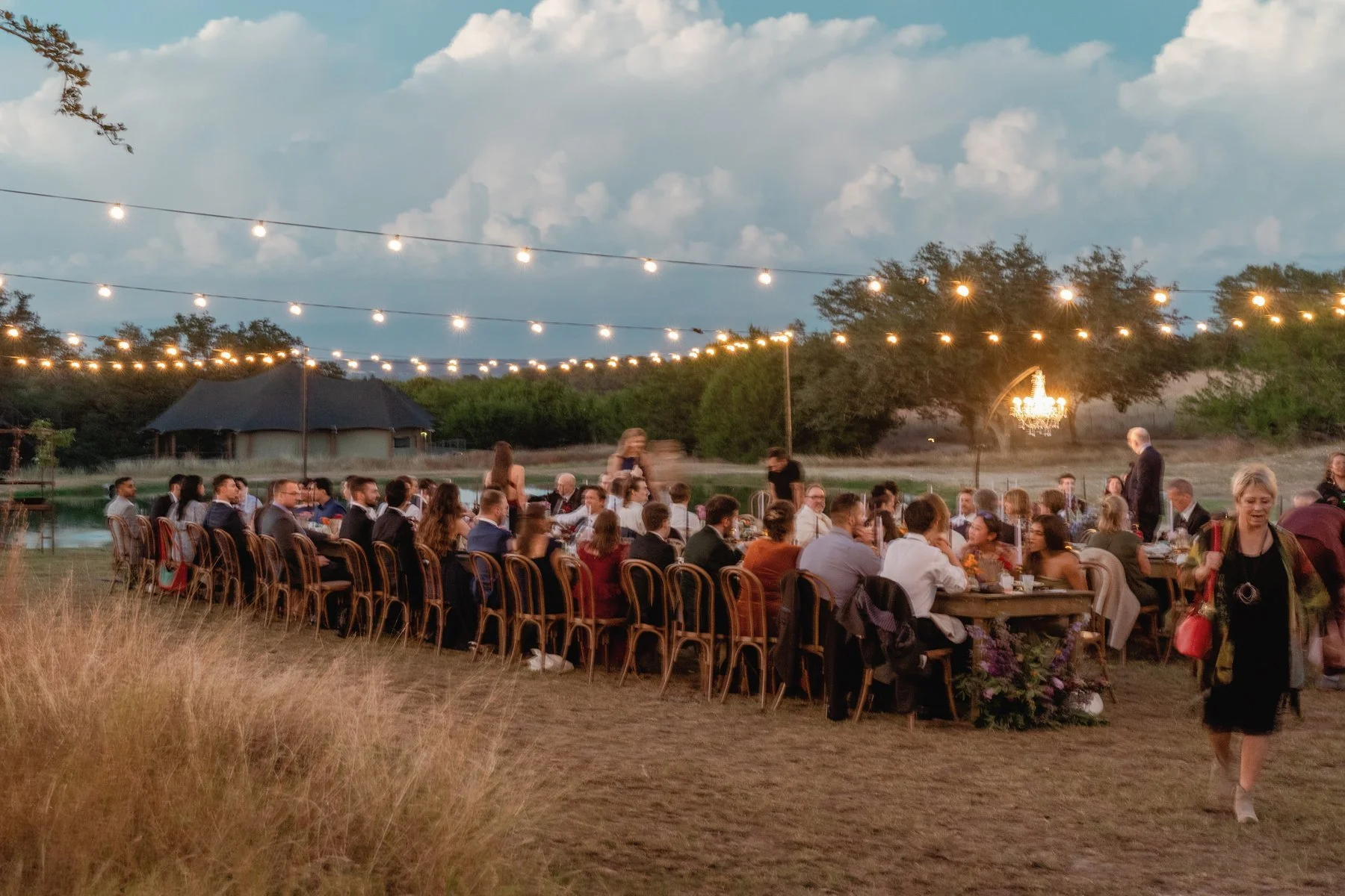 Wide view of the wedding dinner outdoors under string lights at dusk.
