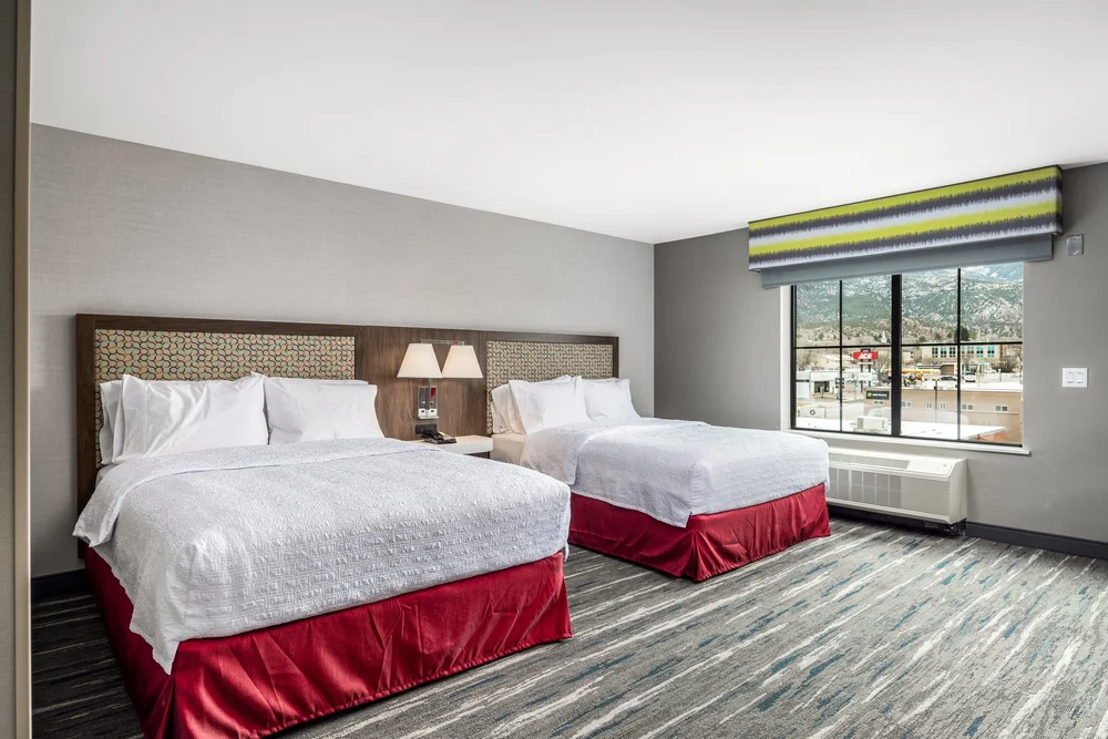 Guest room with two beds, white linens, red bed skirts and a window-side seating area.