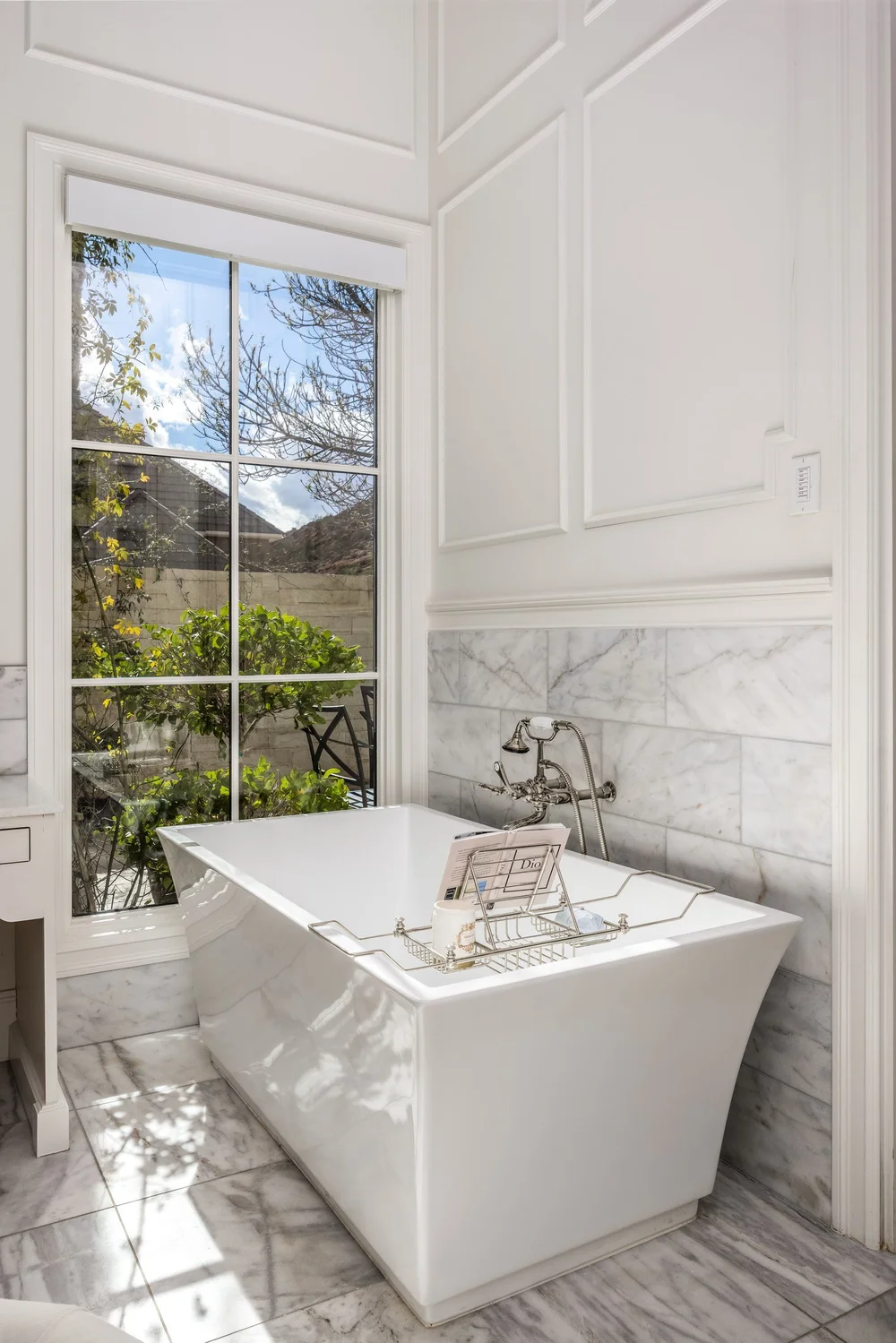 Freestanding bathtub beside a tall window and marble walls