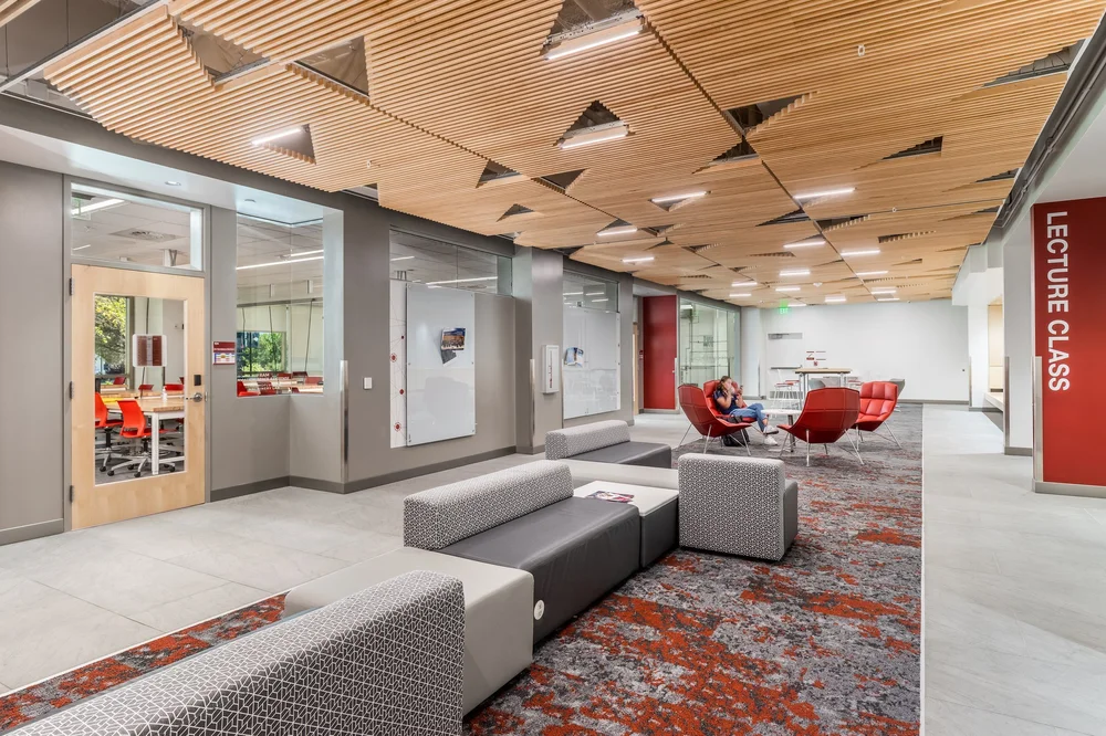 Study lounge with gray seating, red chairs and angular timber ceiling panels.