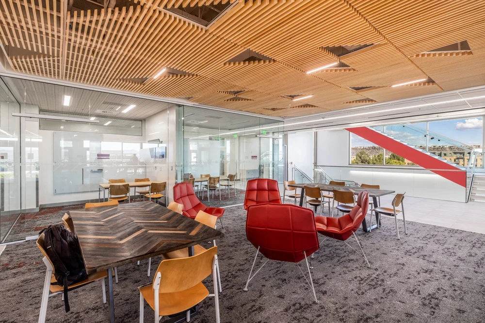 Shared study area beneath a geometric timber ceiling, with red lounge chairs and a glass-walled stair.
