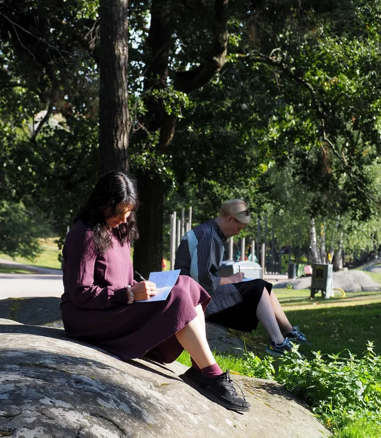 Two people sit on a rock in a park in Helsinki. Under the shade of the trees, they are writing onto a white card - a worksheet from the forests workshop. The sun is dappled through the tree canopy, and there is grass by the edge of the rock.