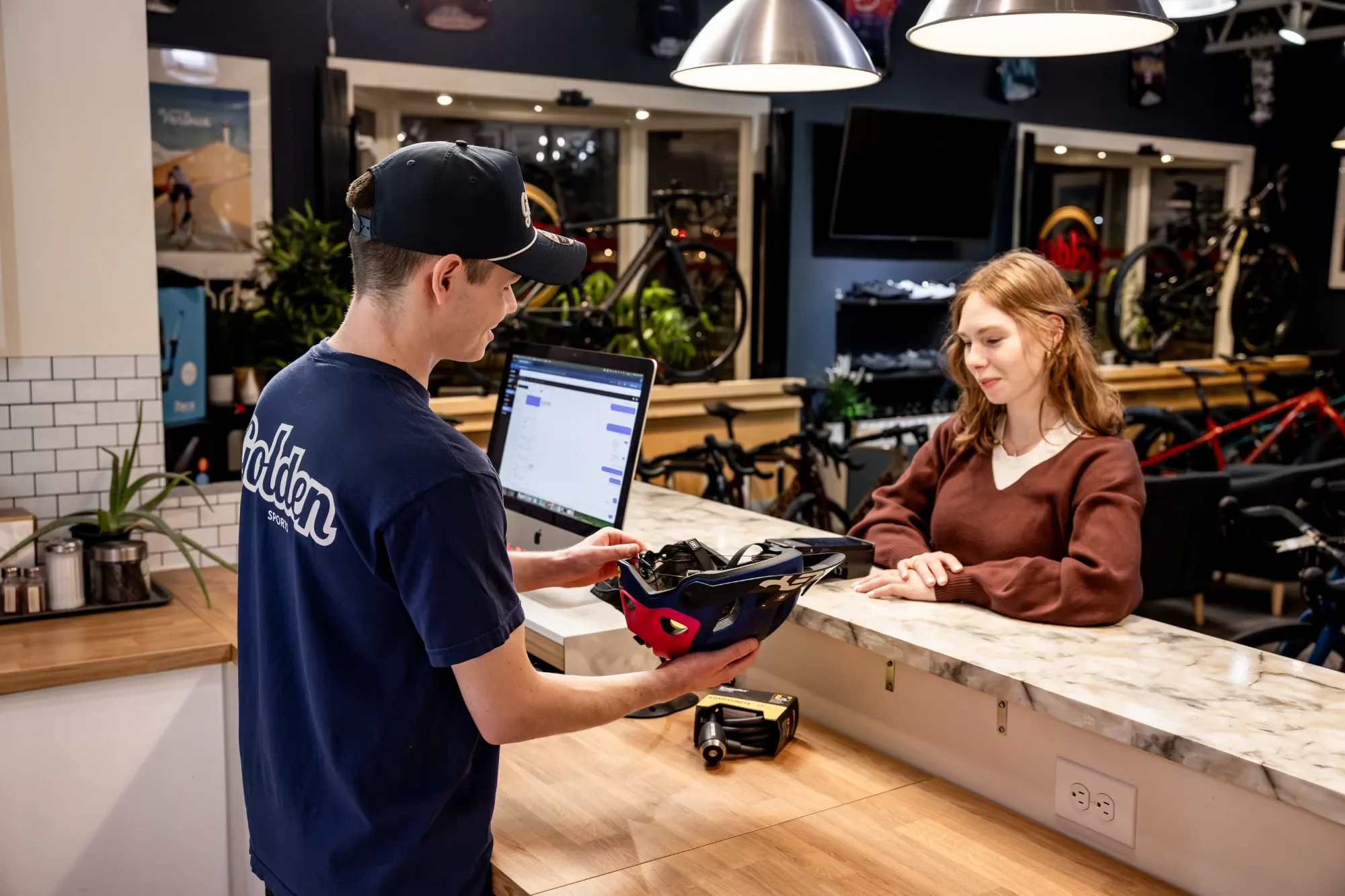 Golden Sports team member helping a customer with a helmet at the counter, with Ikeono open on the computer