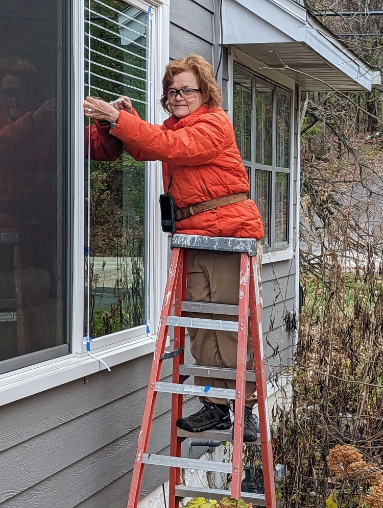 A woman on a ladder applies tape to a window that will help deter birds from colliding with it. The tape is peeled back and leaves a grid of unobtrusive dots.