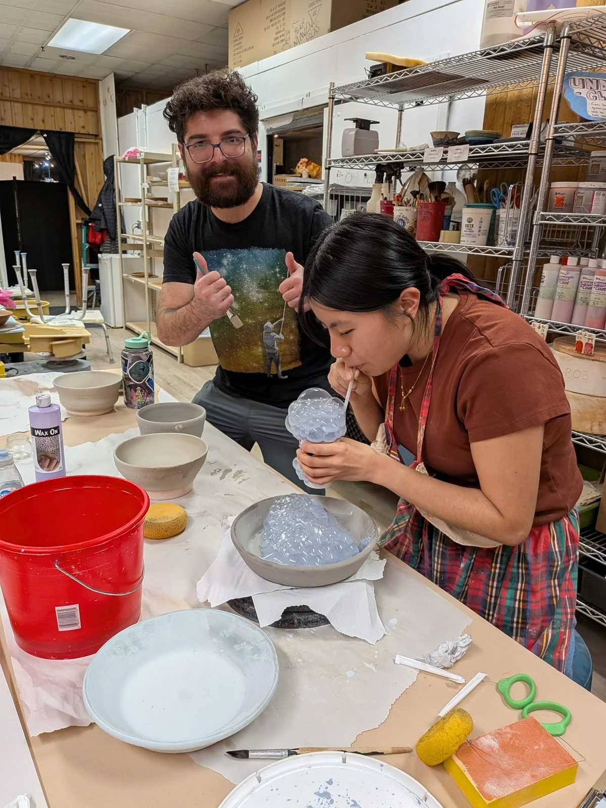 A student blows bubble glaze onto her piece while the instructor gives a thumbs up