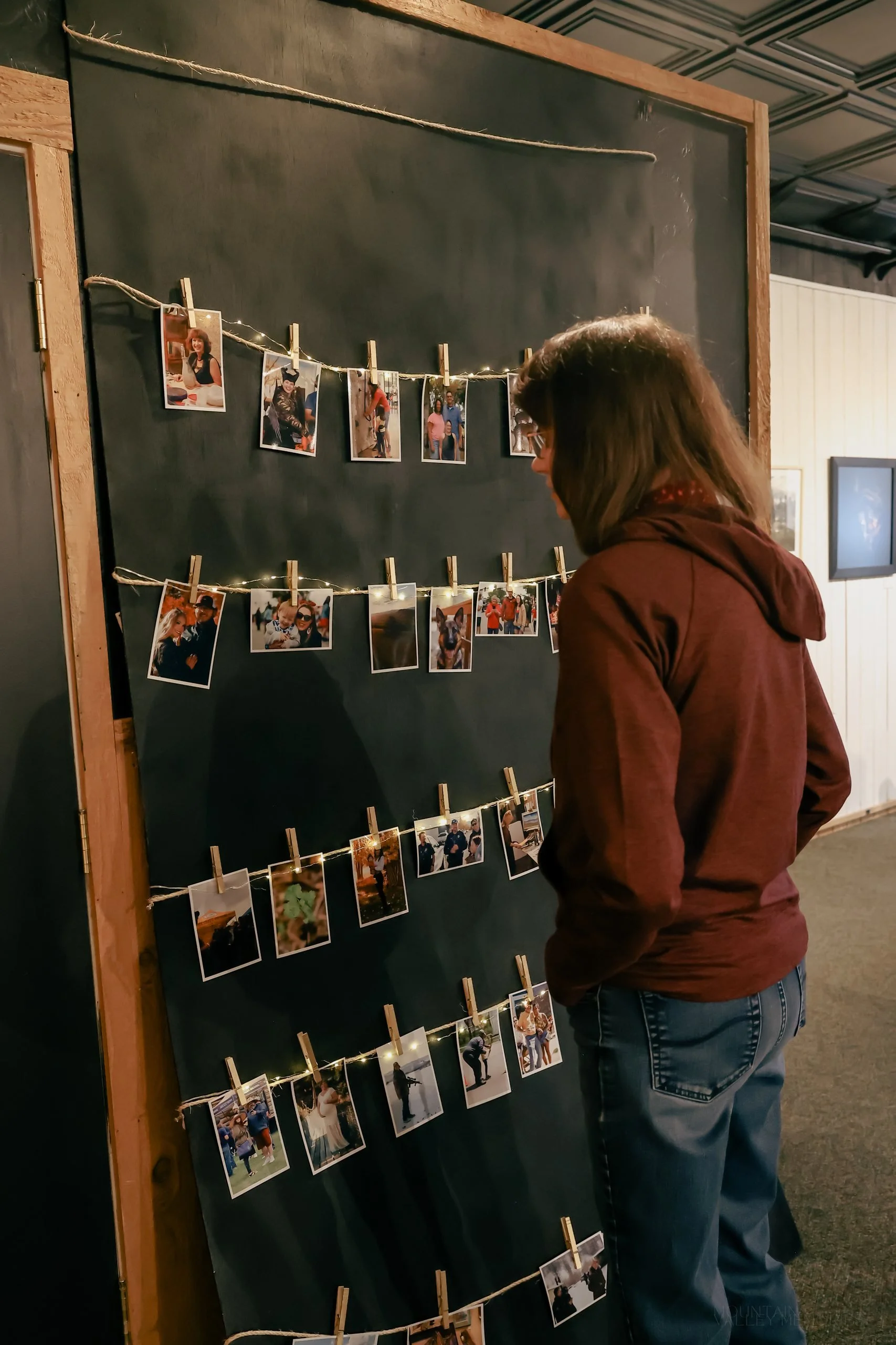 A person in a maroon hoodie looking at snapshots pegged to lines across a chalkboard