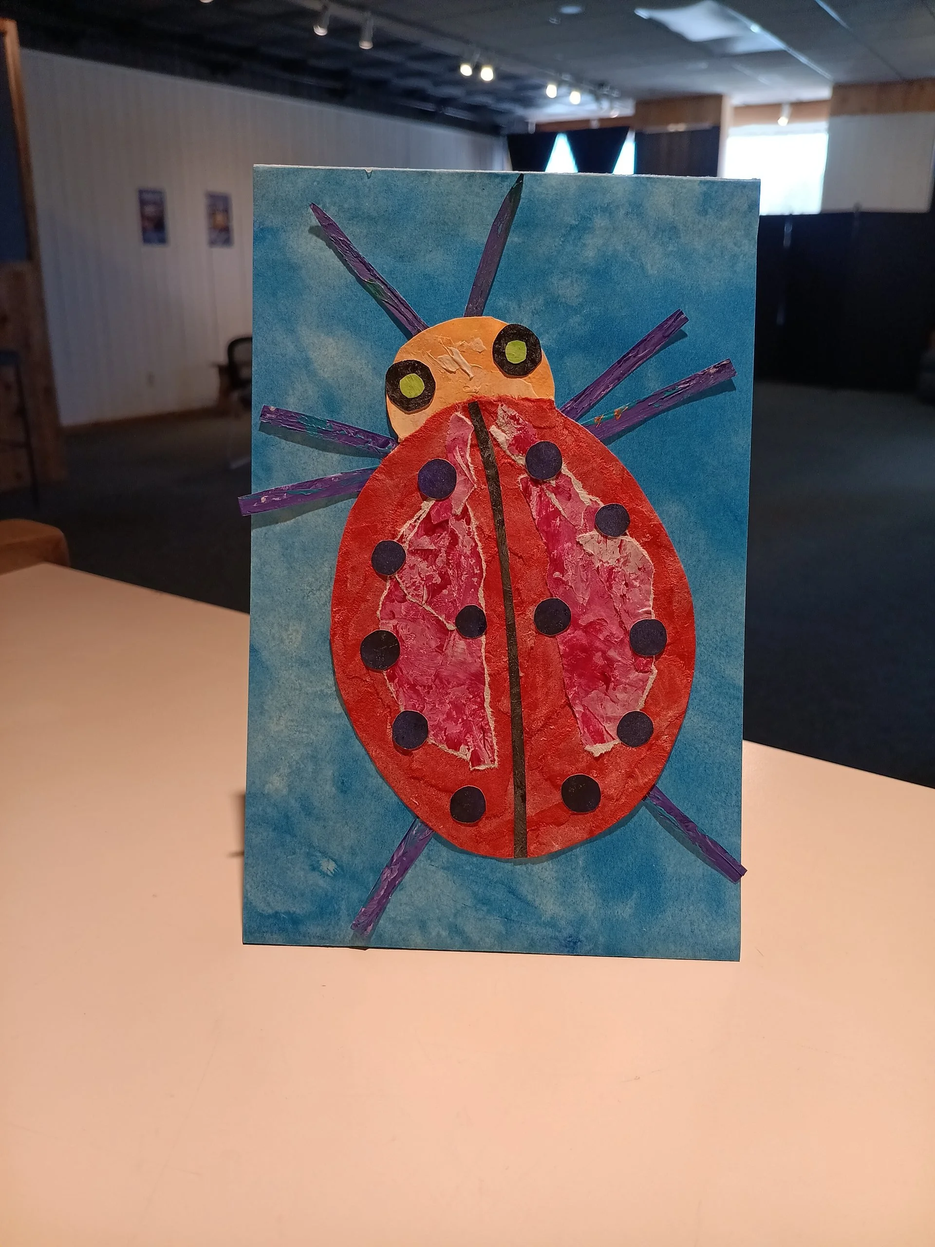 A child's collage of a red ladybug on blue paper standing upright in the gallery