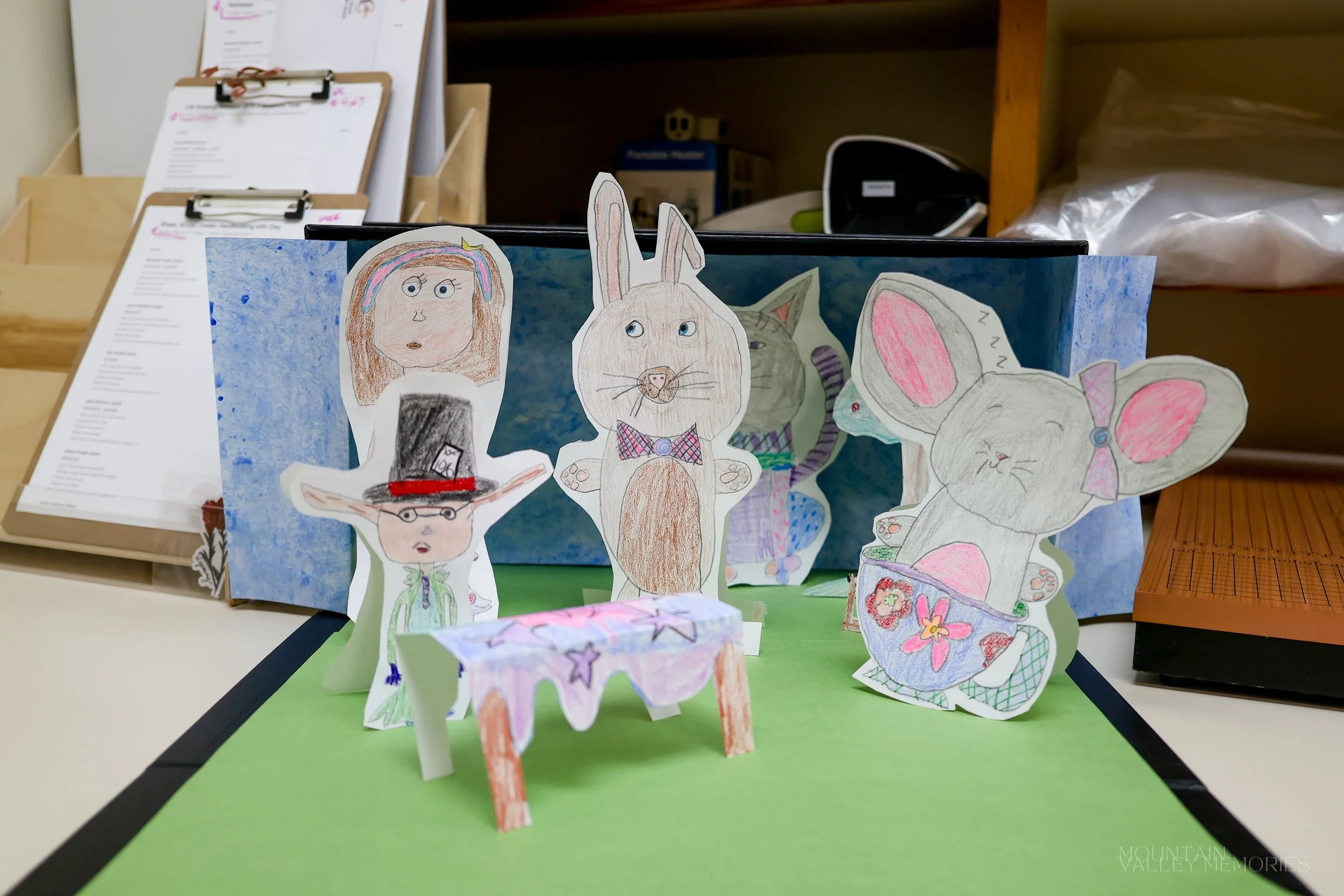 A paper diorama of hand-drawn rabbit and figure cutouts standing around a small table
