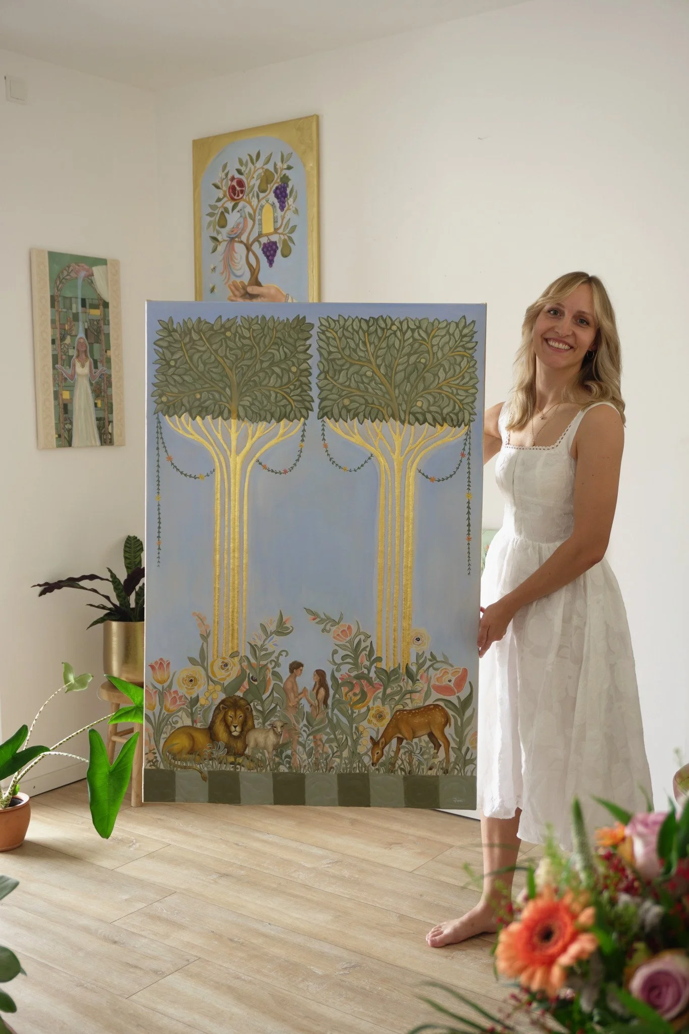 Natalie Ingerle steht im weißen Kleid in ihrem Atelier neben dem großen Gemälde Adam und Eva mit den zwei goldstämmigen Bäumen und hält es am rechten Bildrand