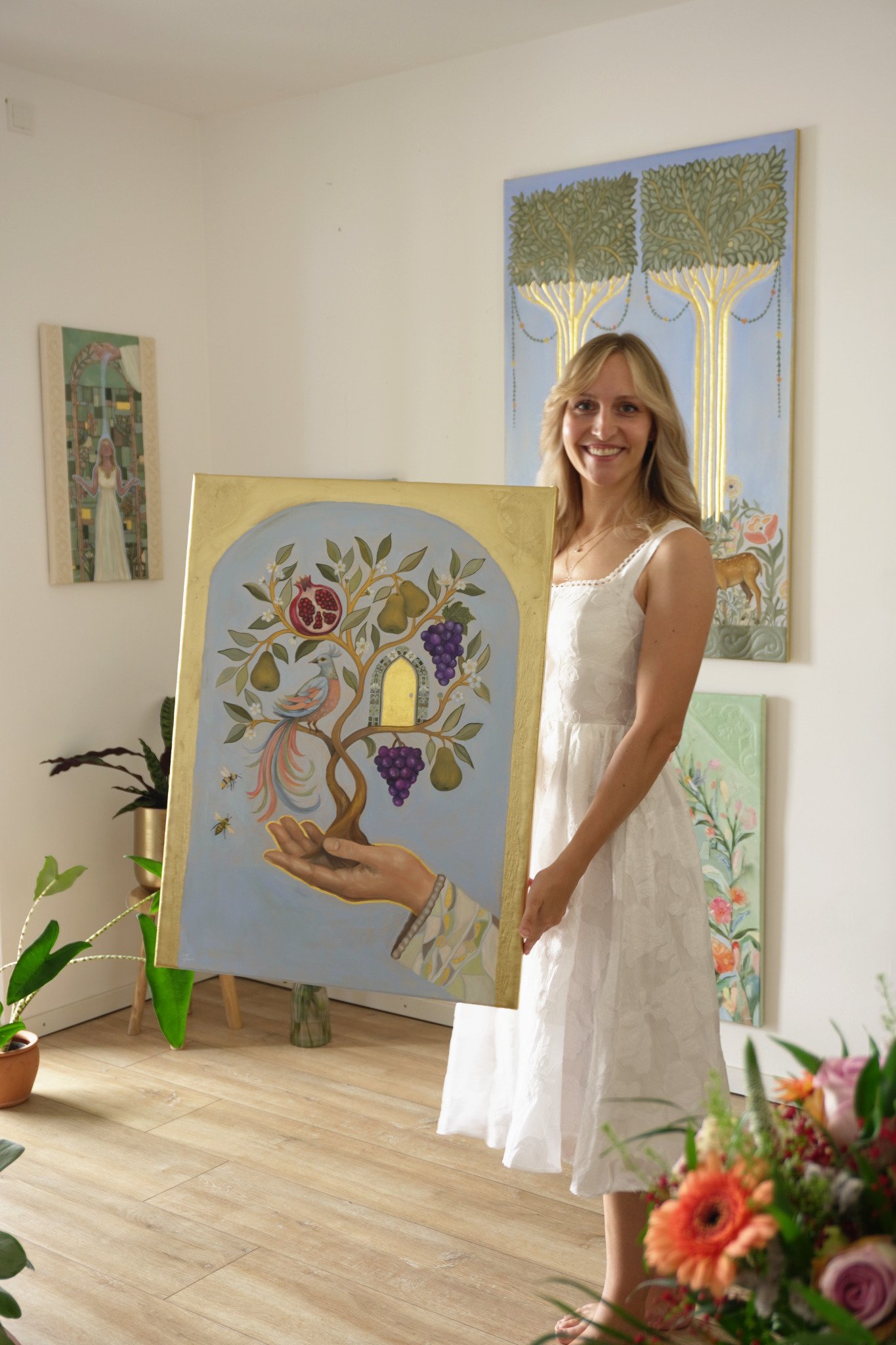Natalie Ingerle hält in ihrem Atelier das goldgerahmte Gemälde Baum des Lebens am Rahmenrand, auf dem ein Fruchtbaum aus einer offenen Hand wächst