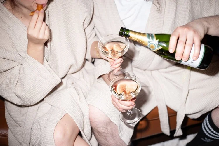 Black and white photo of a couple sitting in robes, champagne being poured for them, styled for a lifestyle couples photoshoot.