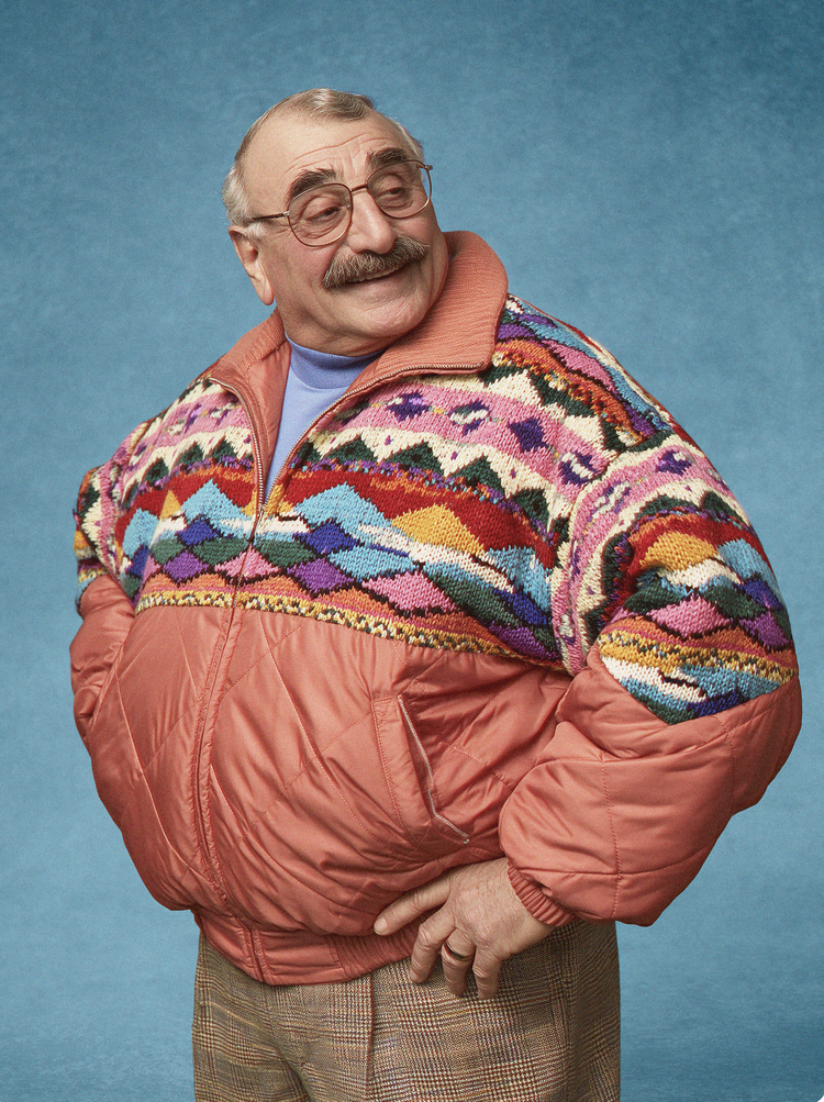 A mustached man in glasses and a patterned ski jacket smiles to himself