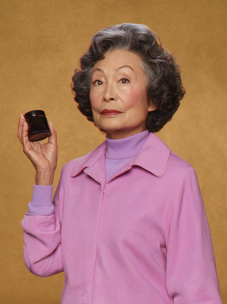 A silver-haired woman in a pink jacket holds up a small jar