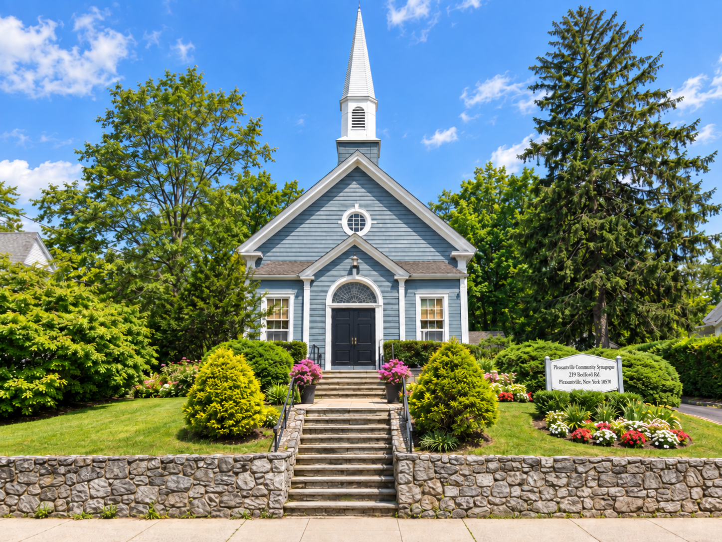 The Pleasantville Community Synagogue from Bedford Road: a blue clapboard sanctuary with a white steeple, stone steps climbing from the sidewalk, and a sign reading Pleasantville Community Synagogue, 219 Bedford Rd., Pleasantville, New York 10570.